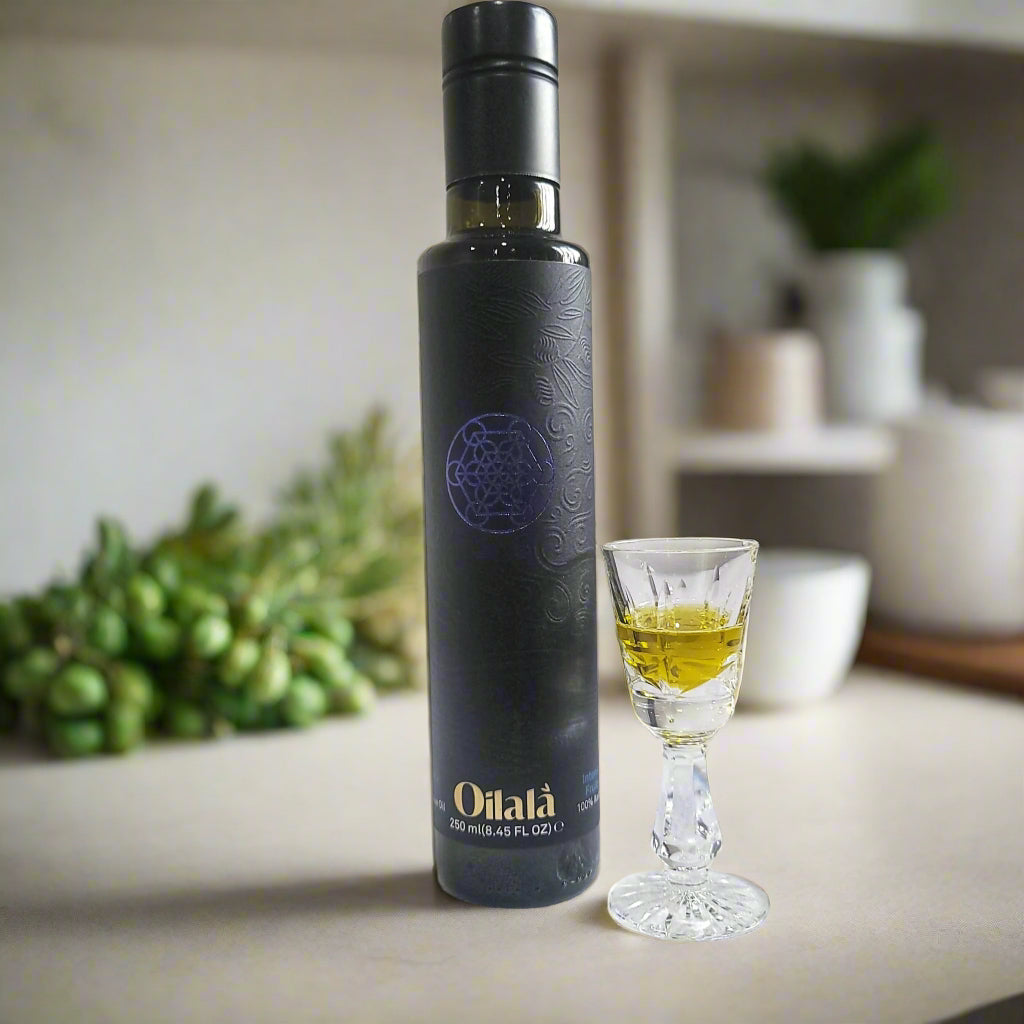A dark glass bottle of Intosso extra virgin olive oil with a label, next to a small glass with a pouring of olive oil, set on a kitchen counter with green olives in the background.