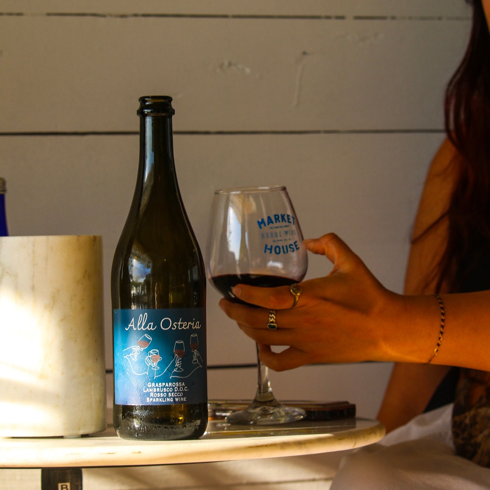 Person holding a glass of wine next to a bottle labeled 'Alla Osteria' on a wooden surface.